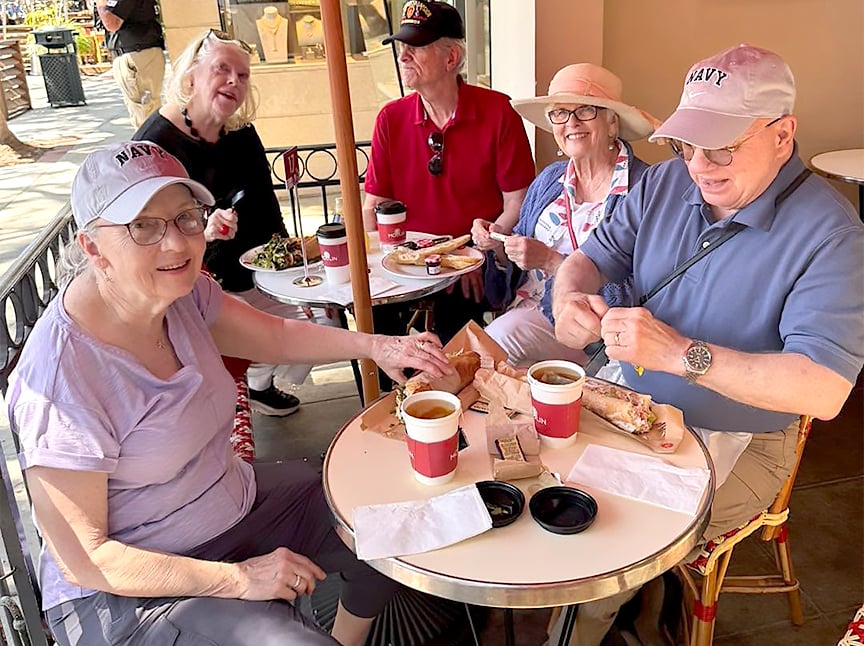 Five older adults sit around a small outdoor café table, smiling and enjoying food and drinks. Three wear light-colored NAVY hats, and everyone appears relaxed and happy in the sunny setting.