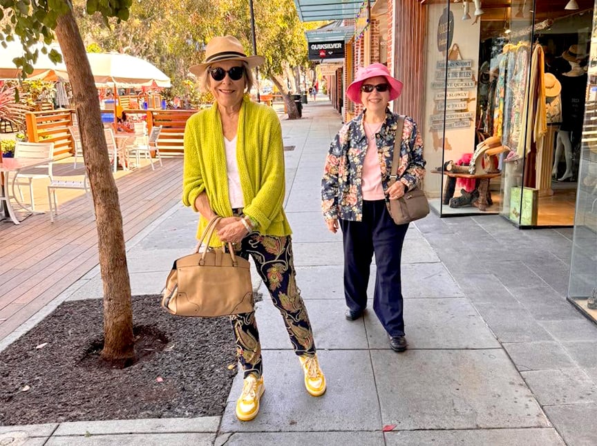 Two older women walk on a sunny outdoor shopping street. One wears a yellow sweater, hat, and patterned pants; the other wears a pink hat and floral jacket. Both are smiling and carrying bags.