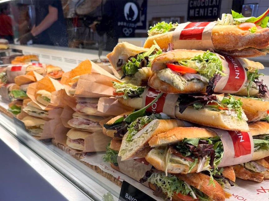 A display case filled with assorted fresh baguette sandwiches, wrapped in paper and packed with lettuce, tomato, cheese, and deli meats in a bakery or café setting.