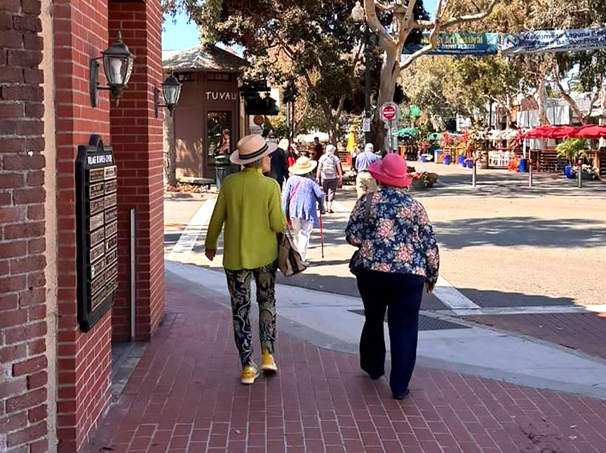 Two women wearing colorful hats and clothes walk down a brick sidewalk in a small town. Other people stroll ahead, and trees, shops, and street signs are visible in the background under a sunny sky.