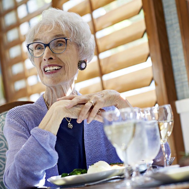 Smiling elderly woman with white hair and glasses sits at a table with food and drinks, wearing a lavender sweater and jewelry, in a sunlit room with wooden shutters.