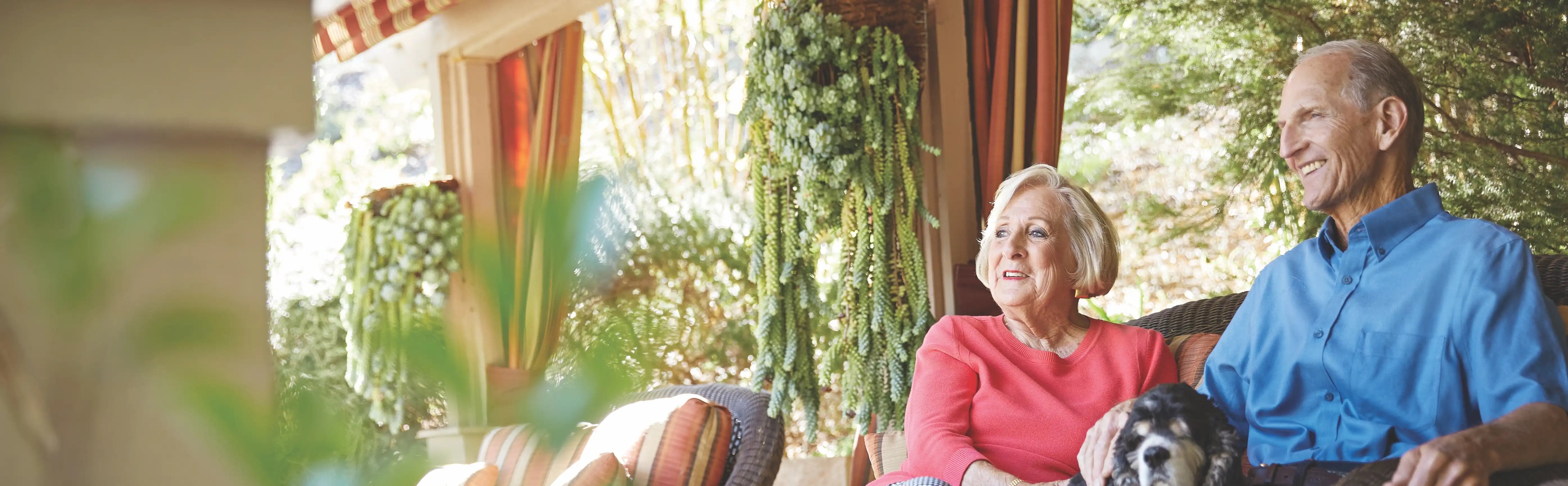 An older woman and man sit together on an outdoor patio, smiling and relaxing. The woman has a small dog on her lap. Green plants and sunlight fill the background, creating a peaceful atmosphere.