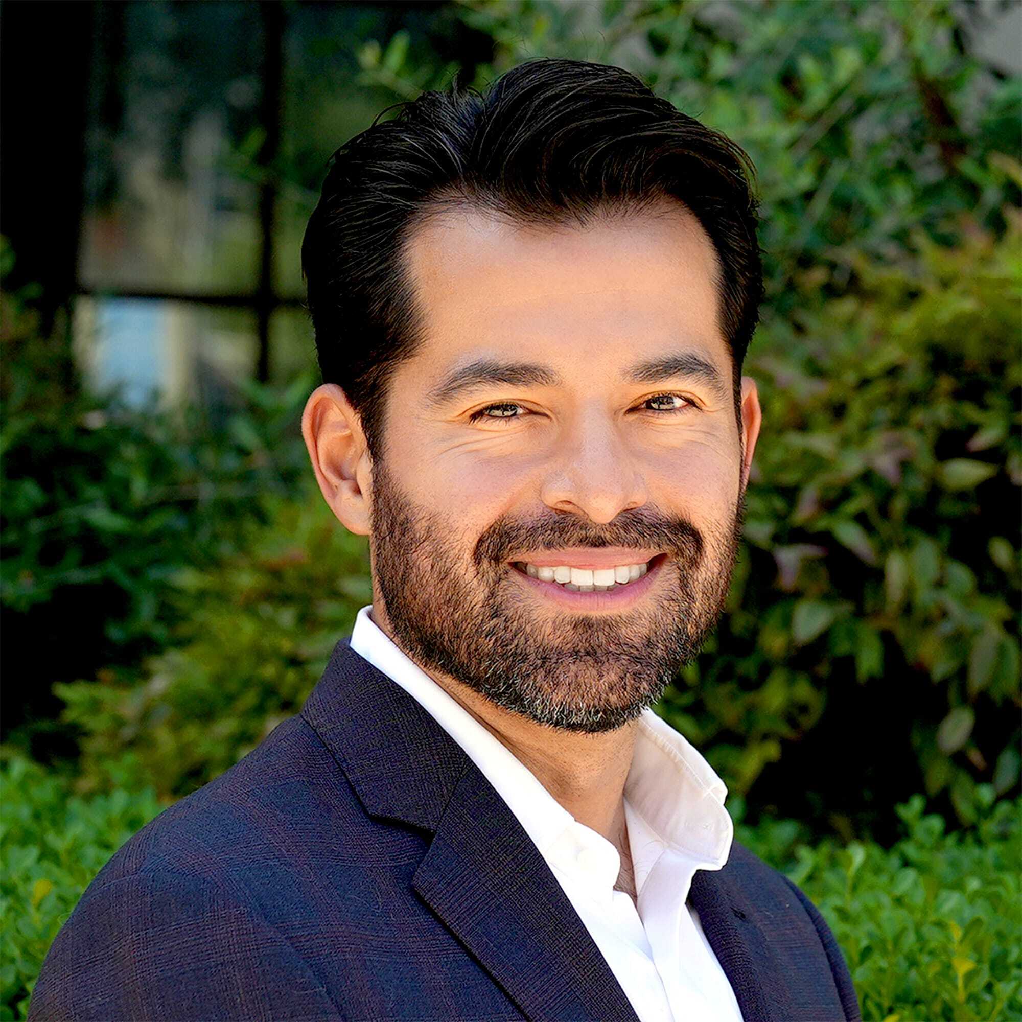 A man with dark hair and a beard, wearing a navy blazer and white shirt, smiles at the camera while standing outdoors with greenery in the background.