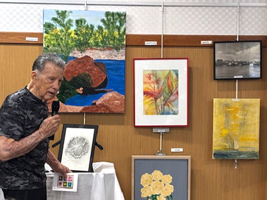 An elderly man speaks into a microphone while standing next to a display of various artworks, including paintings of landscapes, flowers, and a sailboat, in an indoor exhibition setting.