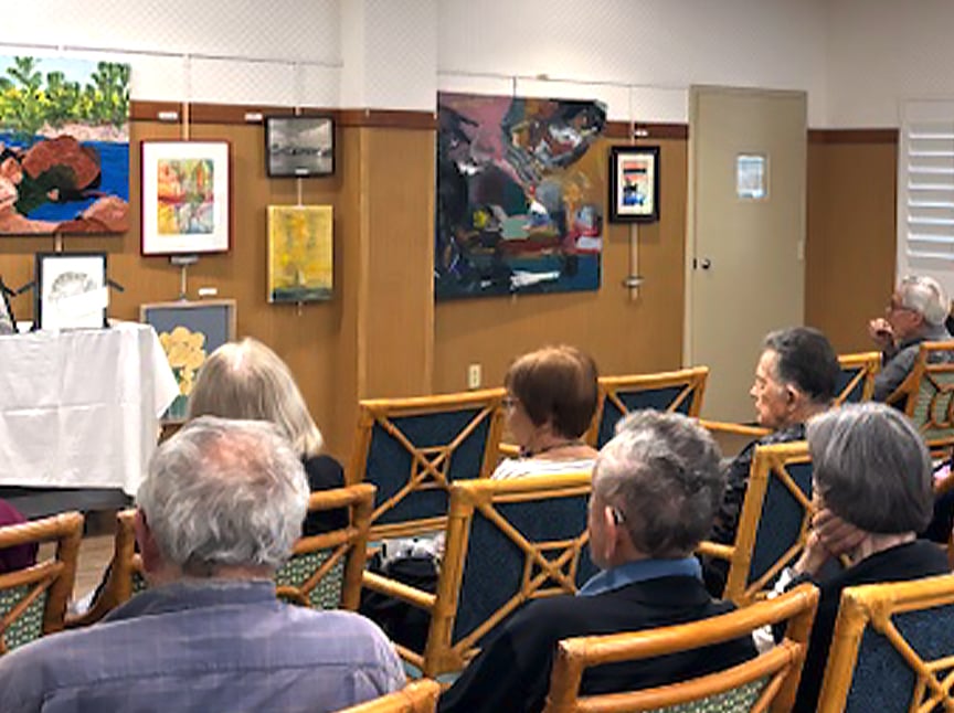 Several older adults sit in chairs facing a display of colorful artwork in a gallery or community room, listening attentively to an unseen speaker. Paintings and drawings are hung on the walls.