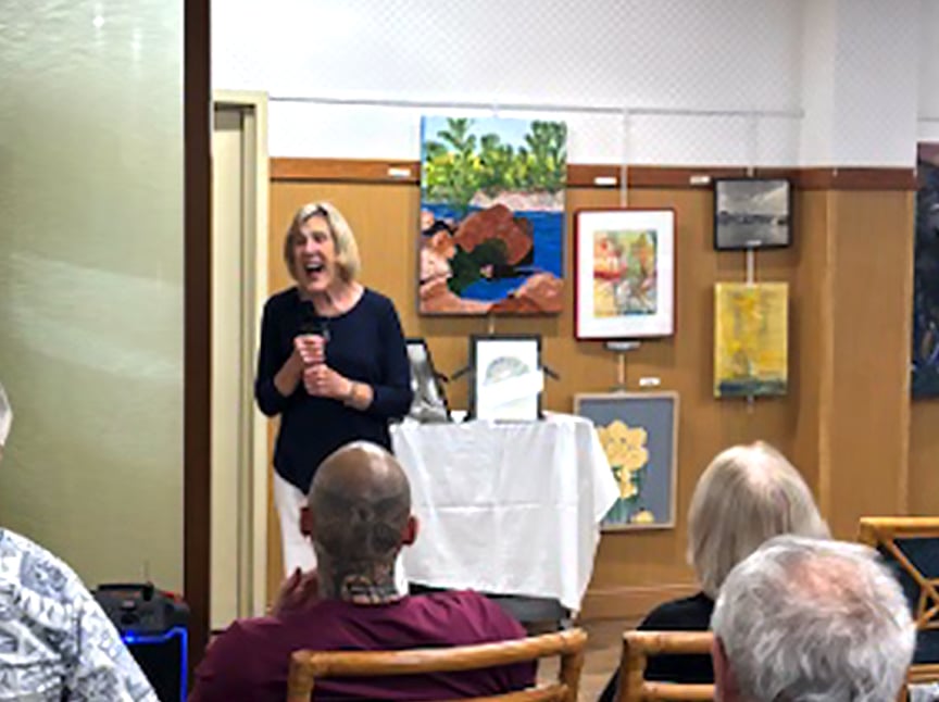 A woman stands speaking in front of an audience in a room decorated with various colorful framed artworks on the walls. She appears to be smiling or laughing. Several people are seated and facing her.