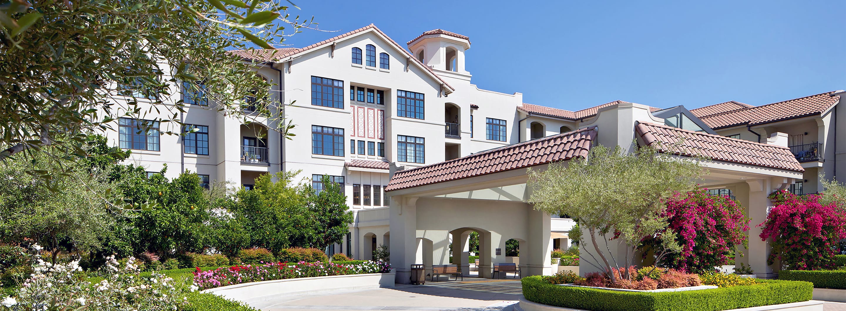 A modern, multi-story building with white stucco walls, large windows, and terracotta roof tiles, surrounded by lush greenery, colorful flowers, and manicured landscaping under a clear blue sky.