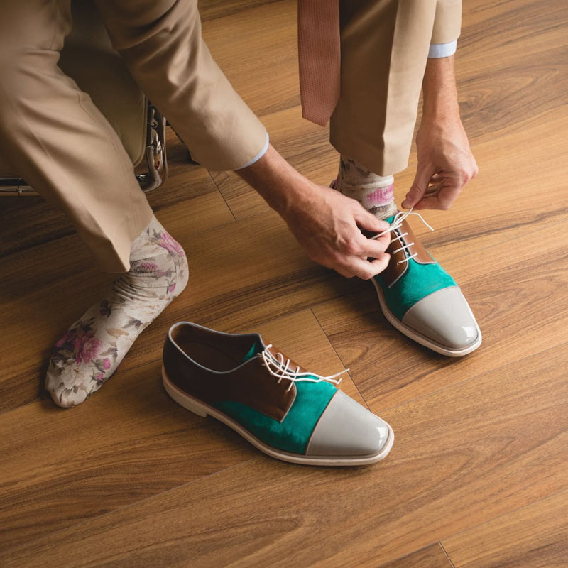 A person wearing a beige suit and floral socks ties the laces on a pair of colorful dress shoes with teal, brown, and gray sections, sitting on a wooden floor.