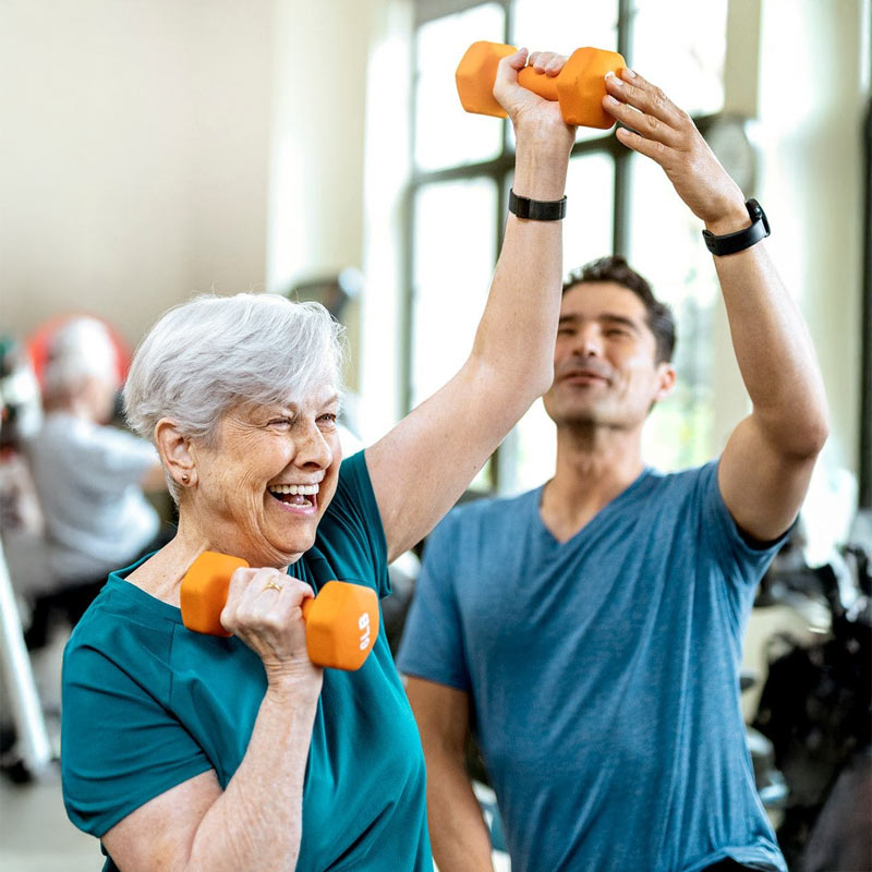 An older woman smiles while lifting orange dumbbells with the assistance of a younger male trainer in a bright gym setting.