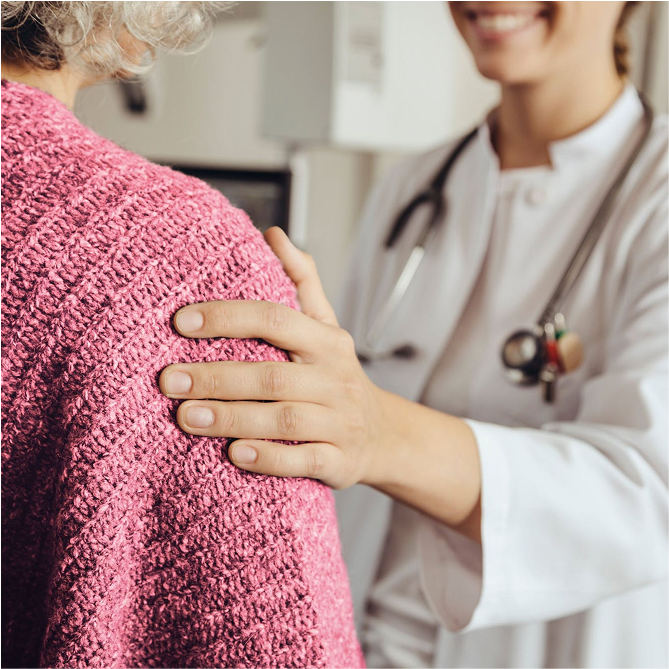 A doctor with a stethoscope gently rests a hand on a patients shoulder, offering comfort. The patient is wearing a pink knitted shawl, and both are indoors in a medical setting.