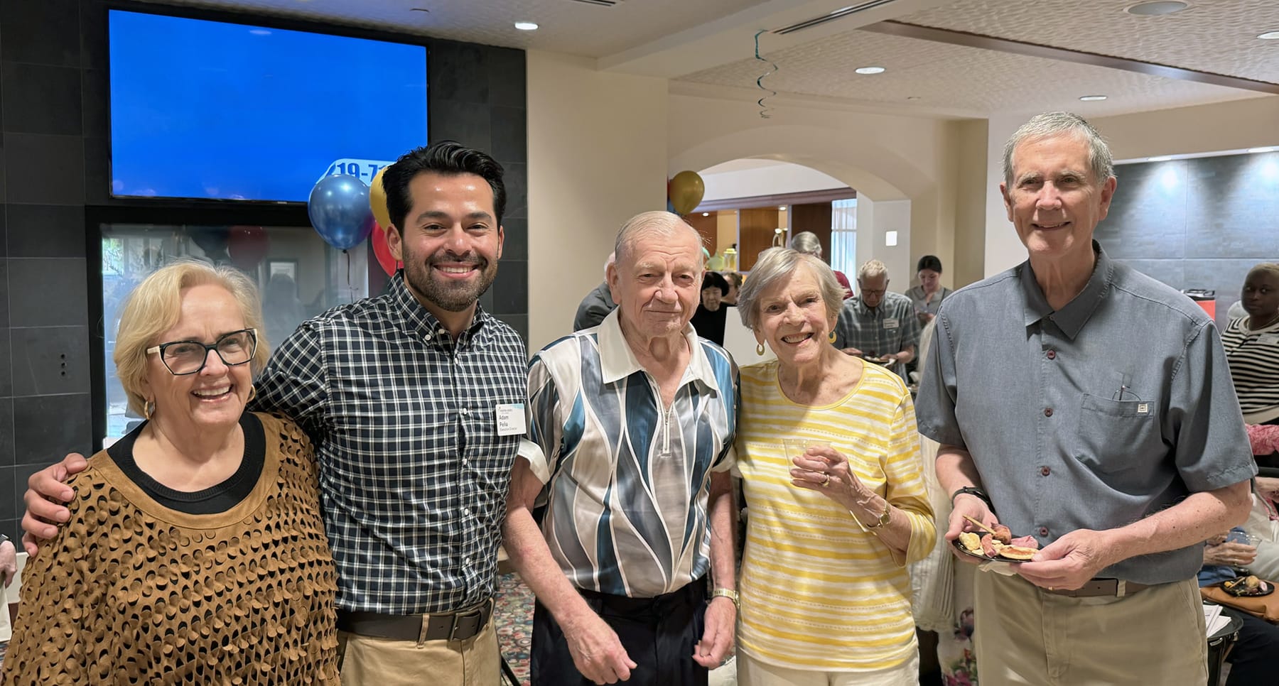Five people, three older men and two older women, stand smiling together at a party with balloons and food. The group poses indoors with others mingling in the background.