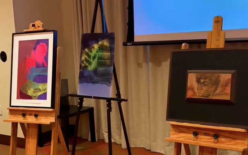 Three easels display artworks: one with vibrant abstract colors and figures, one with a surreal blue-green design, and one with a framed sketch of a face. A projector screen and curtains are in the background.