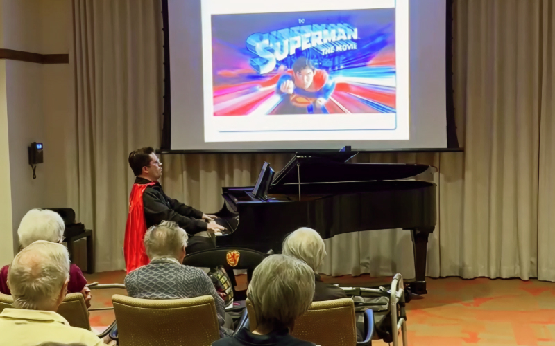 A person wearing a red cape plays a grand piano in front of an audience, with a Superman: The Movie image projected on the screen behind them.