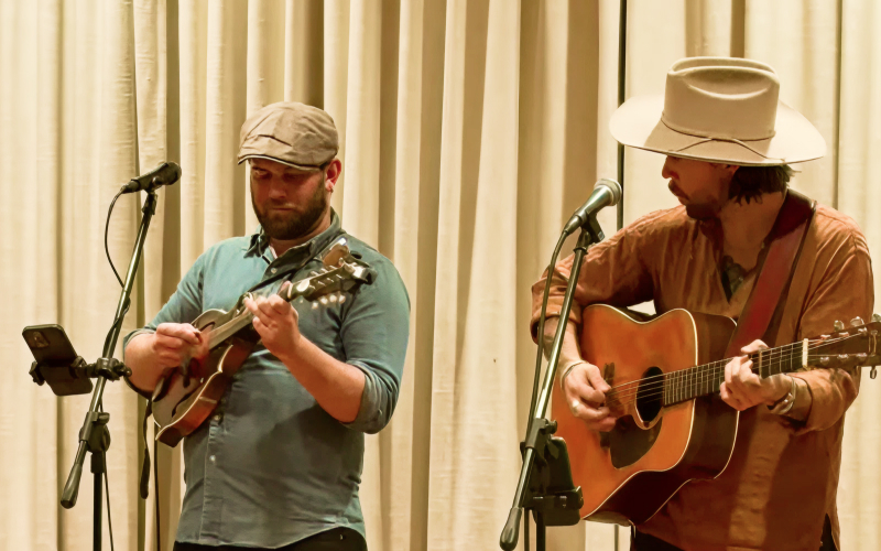 Two musicians perform on stage; one plays a mandolin and wears a flat cap, while the other plays an acoustic guitar and wears a wide-brimmed hat. Both stand in front of microphones and a curtain backdrop.