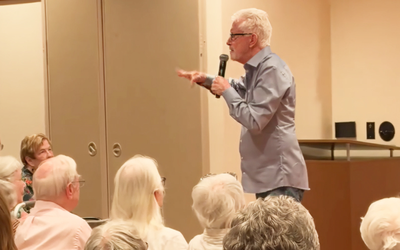 An older man with gray hair and glasses speaks into a microphone while addressing an audience of seniors seated in a room.
