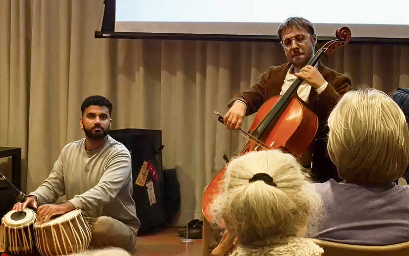 A seated man plays tabla drums while another man stands and plays a cello in front of an audience, with a projection screen and beige curtains in the background.