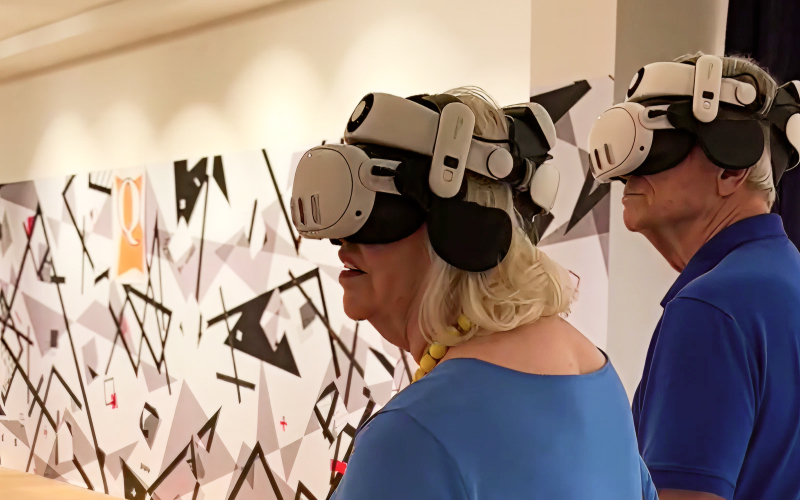 Two older adults wearing virtual reality headsets stand side by side in front of a wall with abstract black and white geometric patterns. They appear focused on their VR experience.