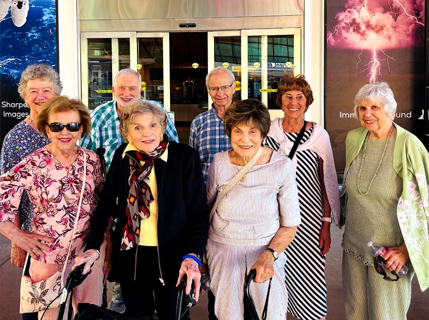 A group of eight smiling older adults, four men and four women, stand together outside a building entrance. Some are wearing sunglasses and light spring clothing. The background has posters of space and a lightning storm.