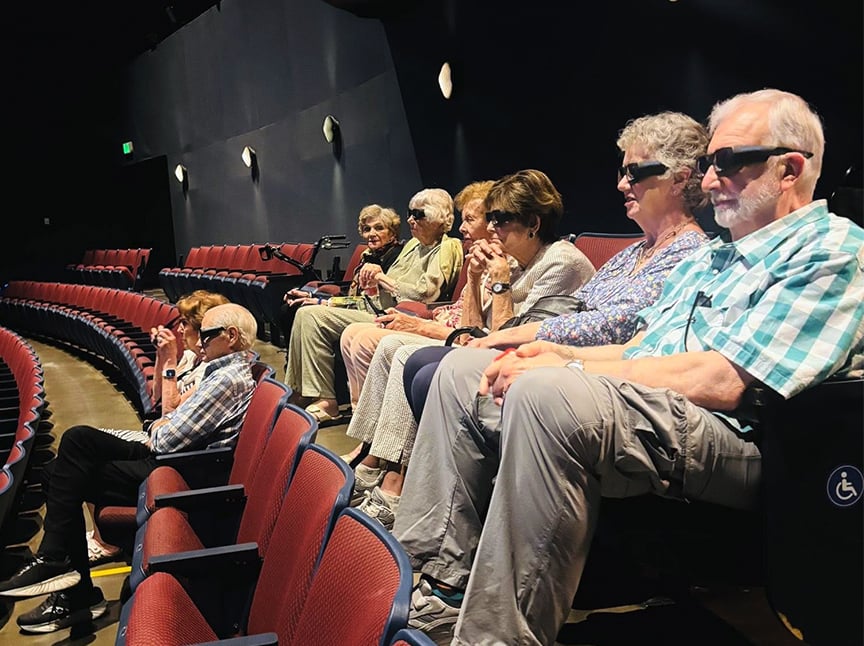 A group of older adults wearing 3D glasses sit in red theater seats, watching a show. Most seats around them are empty, and they appear engaged with the performance.