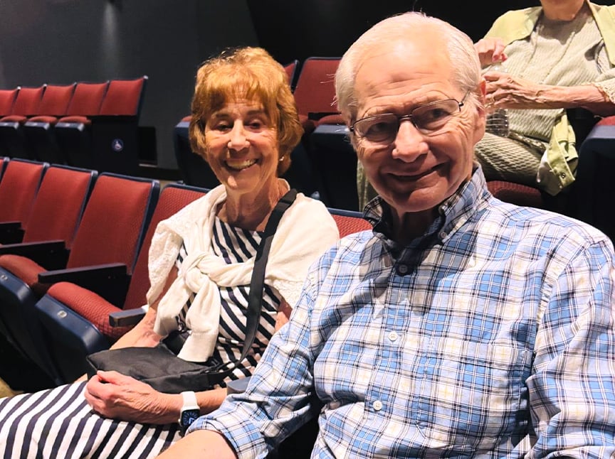 An older woman and man sit together smiling in a theater with red seats. The woman wears a striped dress and white sweater, and the man wears glasses and a blue plaid shirt. Other audience members are in the background.