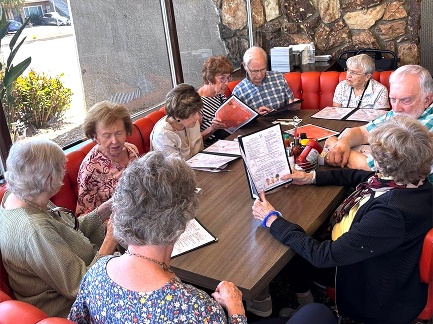 Eight elderly people sit around a table in a restaurant booth, looking at menus and talking. The group is seated on red cushioned benches, with sunlight coming through large windows beside them.