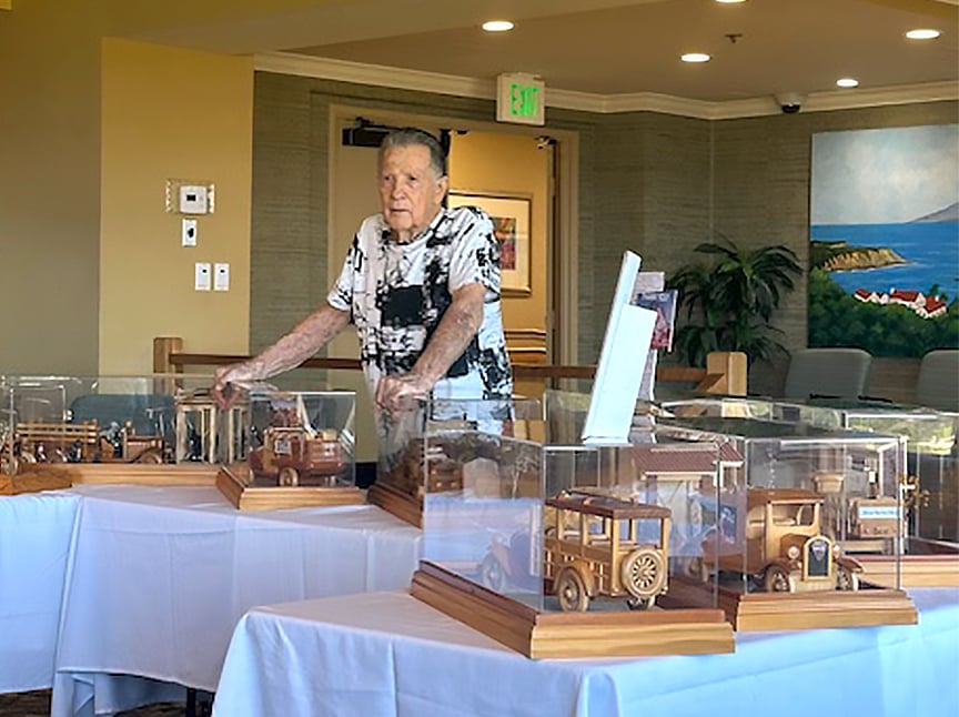 An elderly man stands behind tables displaying detailed wooden model cars in glass cases, inside a well-lit room with paintings, plants, and chairs in the background.