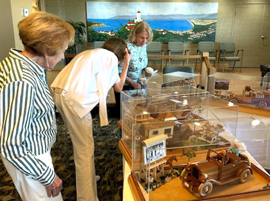 Three older women view detailed miniature dioramas displayed in clear cases on tables. In the background, theres a large painting of a coastal scene with a lighthouse, houses, and ocean. The room has chairs and carpeting.