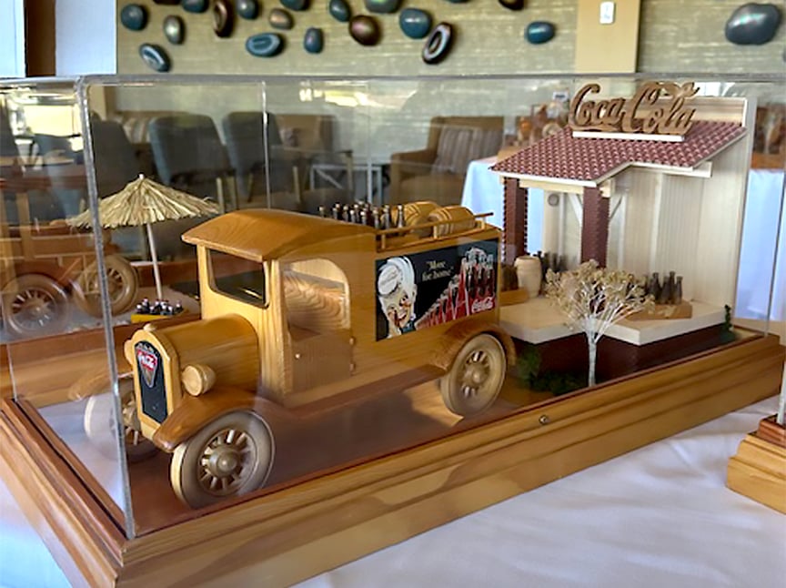 A detailed wooden model of a vintage Coca-Cola delivery truck and storefront is enclosed in a glass display case, featuring tiny soda bottles and decorative elements like trees and an umbrella.