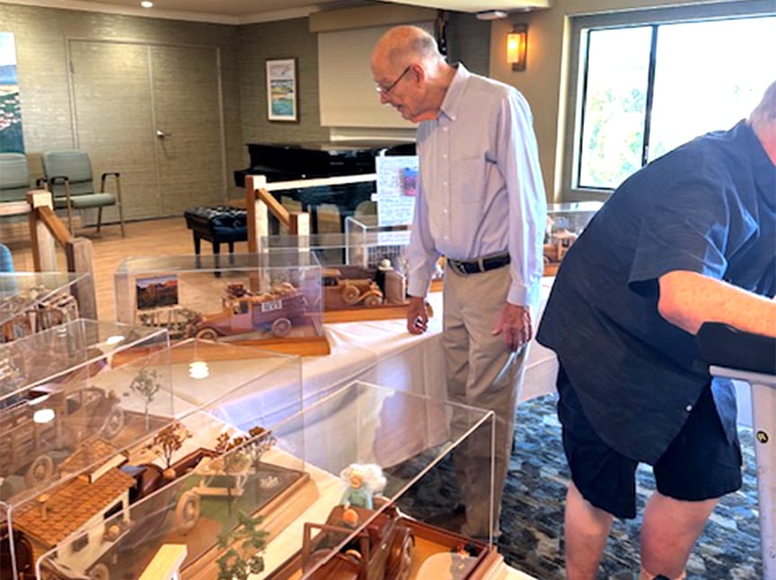An elderly man looks at detailed wooden model cars displayed under clear cases on tables in a well-lit room, while another person leans on a walker nearby.