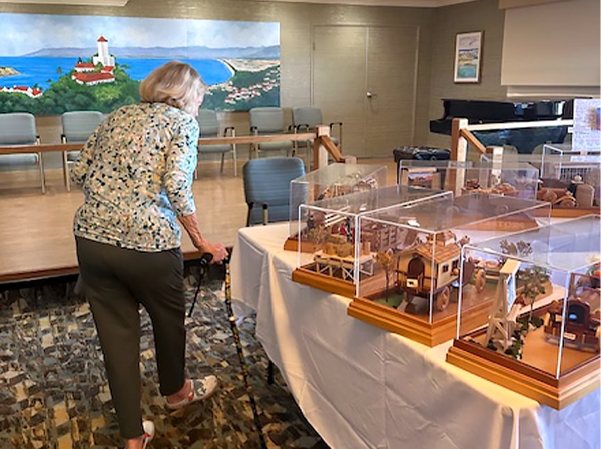 An older woman with a cane looks at several detailed dollhouse miniatures displayed on a table, with a stage, chairs, and a large painting of a coastal scene in the background.