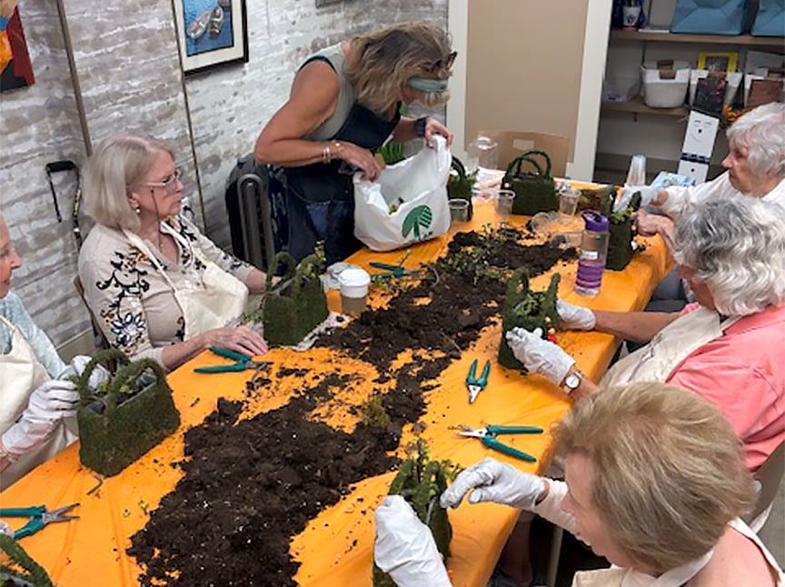 A group of older adults sits around a table covered with soil and gardening tools, working on small plant arrangements. An instructor stands nearby, assisting and providing materials in a bright, indoor setting.