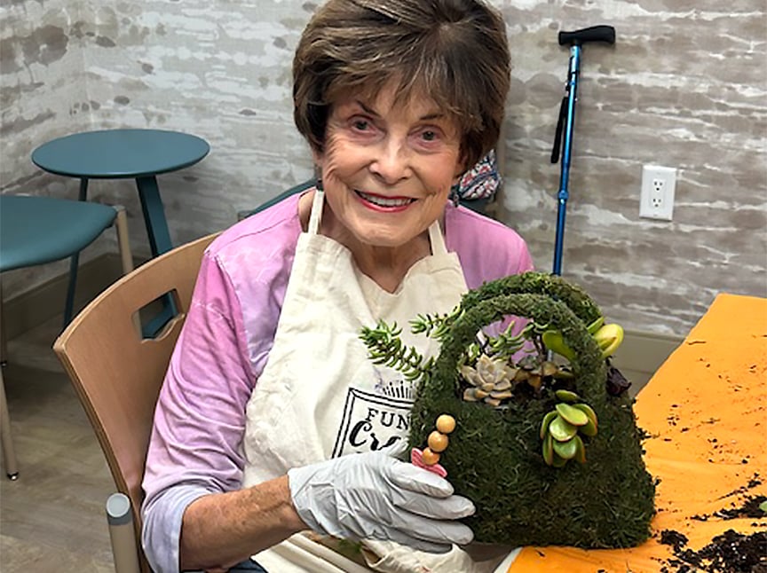 An elderly woman wearing gloves and a white apron smiles while holding a moss-covered basket decorated with succulents and small ornaments. She is seated at a table with crafting materials.