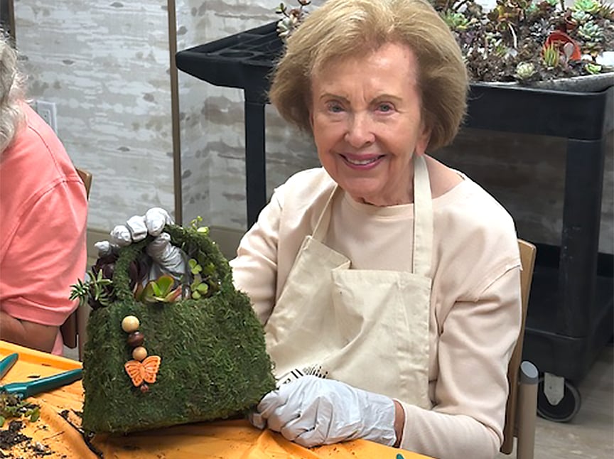 An elderly woman smiles while holding a purse-shaped planter covered in moss and decorated with small succulents, beads, and a butterfly ornament. She wears an apron and gloves, sitting at a craft table.