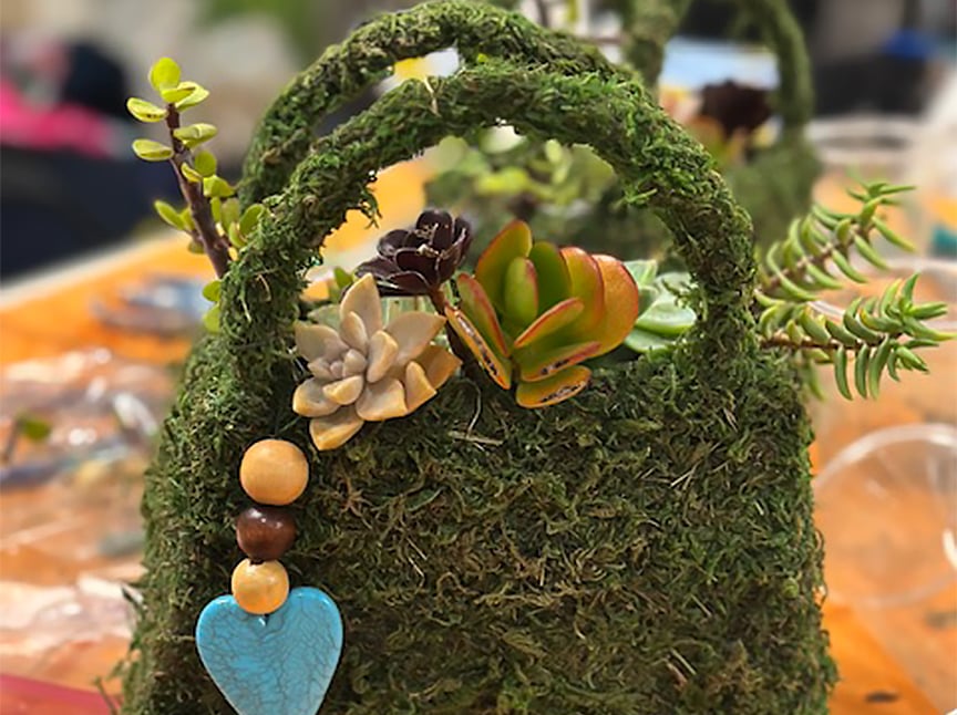 A moss-covered bag decorated with colorful succulents, wooden beads, and a blue heart-shaped charm sits on a table with another similar bag blurred in the background.