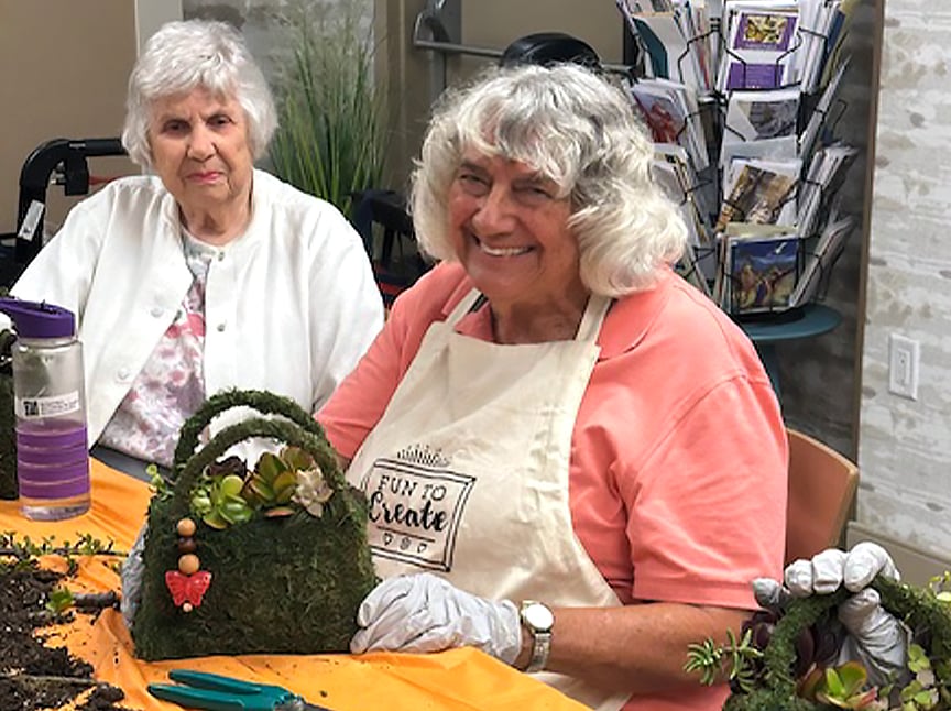 Two elderly women sit at a table crafting with plants. One woman, smiling and wearing an apron and gloves, holds a basket decorated with succulents. The other woman sits beside her, watching. Gardening materials are on the table.