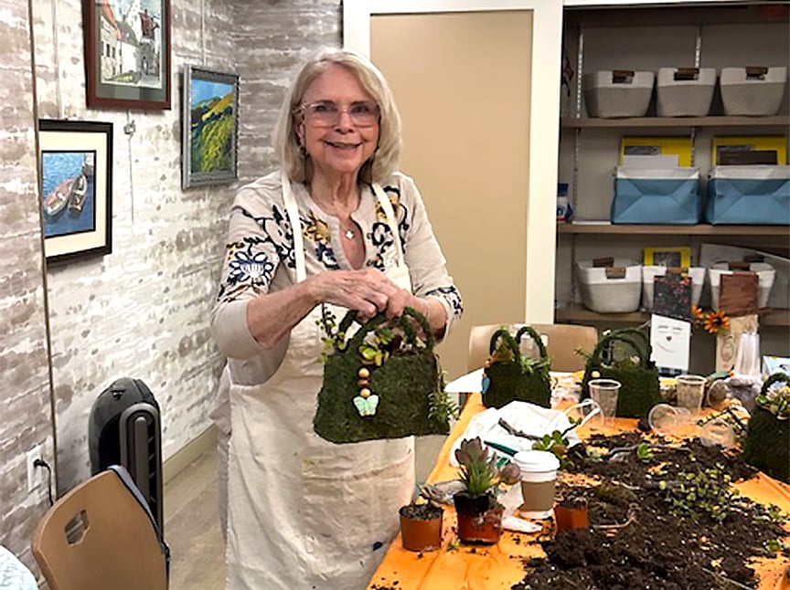 An older woman wearing glasses and an apron smiles while holding a moss-covered decorative basket at a craft table, surrounded by succulents, craft materials, and finished baskets in a cozy, art-filled room.