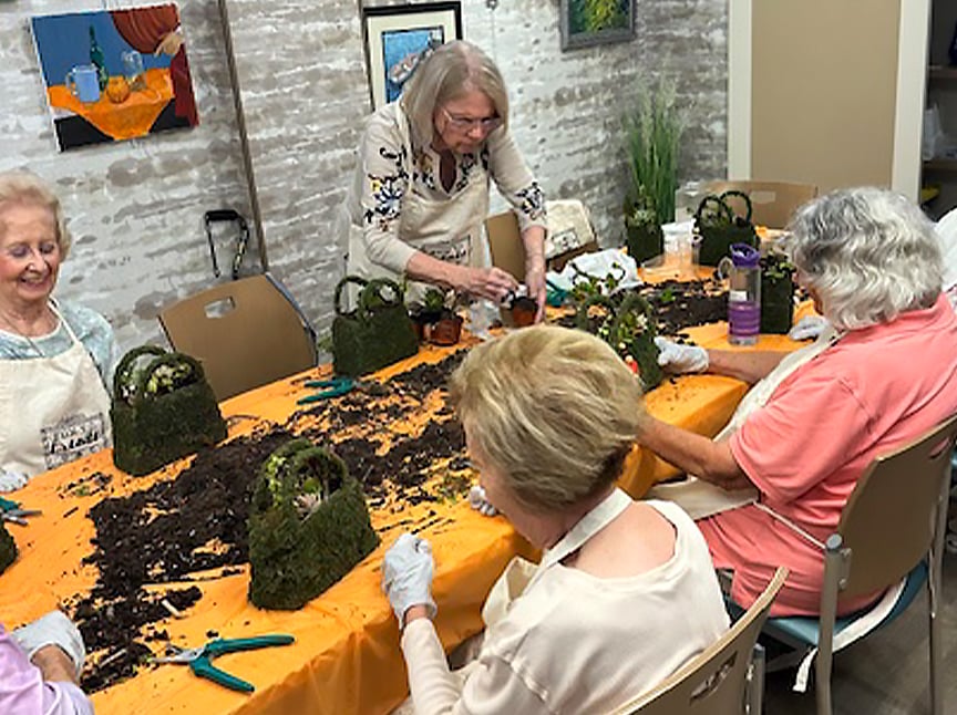 Four older women sit around a table covered with soil and gardening tools, working on planting projects in a bright room with paintings on the wall. They appear engaged and are wearing gloves.