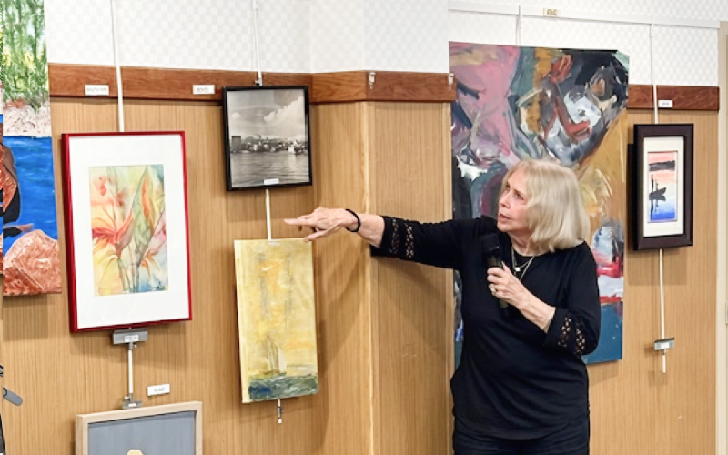 A woman with blonde hair, dressed in black, holds a microphone and gestures toward a gallery wall displaying several colorful paintings and photographs.