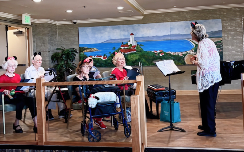 A group of elderly women wearing mouse ears sit with walkers and music sheets, while a woman stands in front conducting. They are indoors, with a seaside painting on the wall behind them.