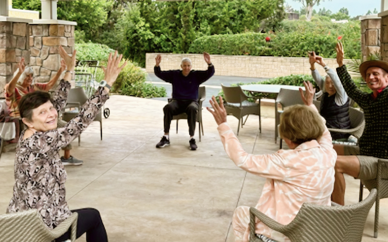 A group of seniors sits in a circle outdoors, raising their arms while participating in a group exercise. The background features greenery and a stone wall. Everyone appears engaged and smiling.