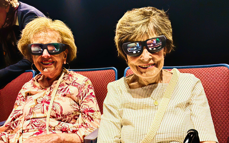 Two older women sit side by side in red theater seats, both smiling and wearing large 3D glasses. Bright lighting highlights their faces and patterned tops against the dark background.