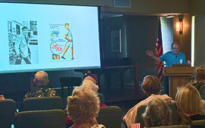 A man stands at a podium giving a presentation to an audience. A projector screen shows two vintage movie posters featuring people and text. The audience sits facing the speaker in a room with an American flag in the background.