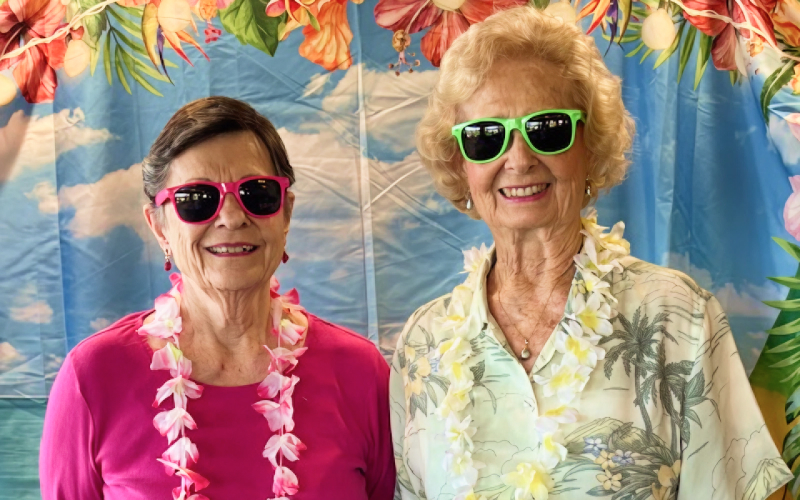Two older women wearing colorful leis and sunglasses stand smiling in front of a tropical-themed backdrop decorated with flowers and palm leaves.