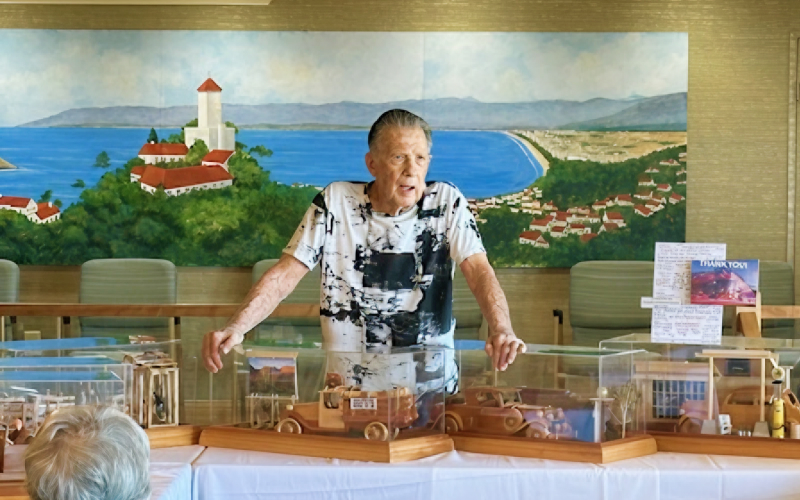 An elderly man stands behind a table displaying handcrafted wooden vehicles in clear cases, speaking to an audience. A mural of a coastal town and ocean is painted on the wall behind him.