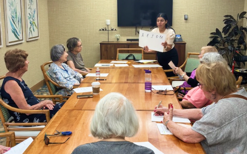 A woman stands at the front of a table holding a sign with letters while several older adults sit around the table, attentively watching and listening. Papers and water bottles are on the table.