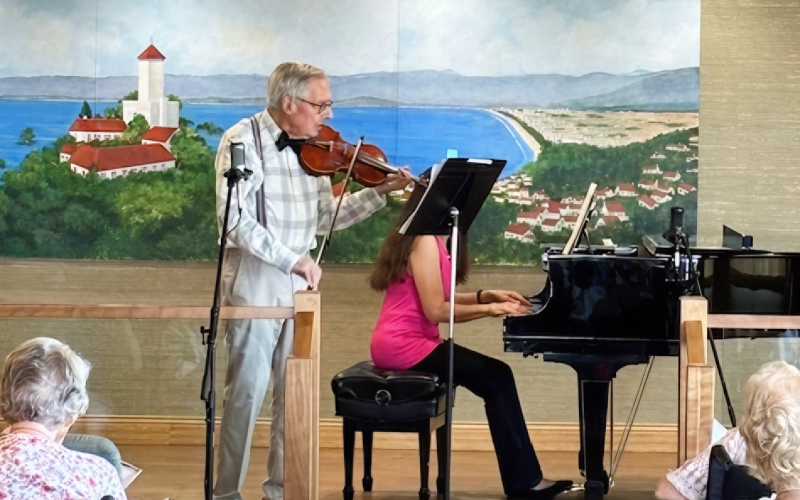 An elderly man plays the violin beside a seated woman in a pink top playing a grand piano. They perform in front of an audience with a scenic mural of a lake and village in the background.