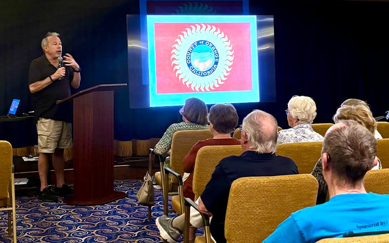 A man speaks at a podium to an audience seated in rows, with a large screen behind him displaying a brightly colored flag with a circular emblem at the center. The setting appears to be an indoor presentation or lecture.