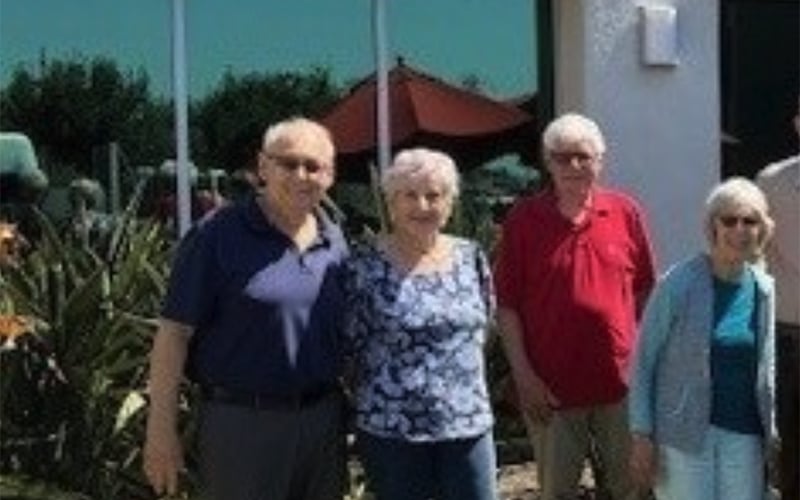 Four older adults stand outdoors in front of a building and plants on a sunny day. Two men and two women are smiling, with a red umbrella and green trees visible in the background.