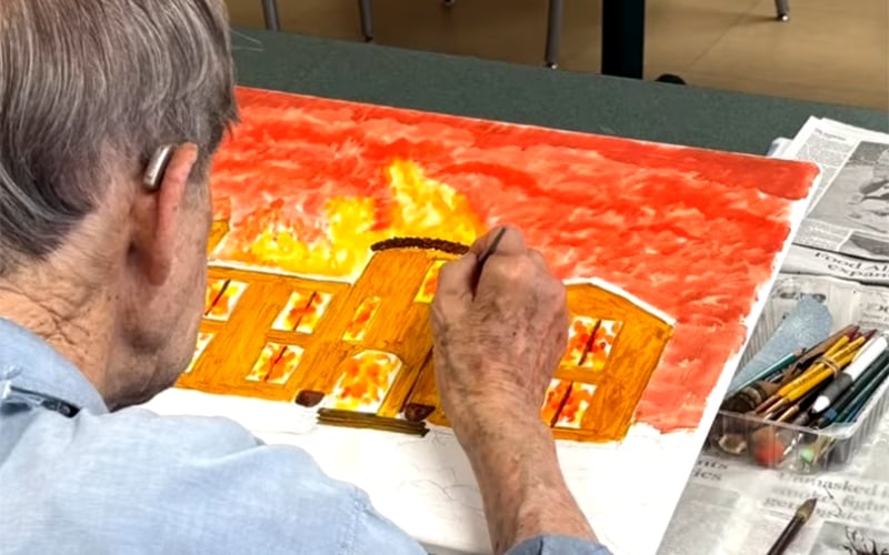 An elderly person with gray hair is painting a picture of a burning building with bright orange flames and smoke, using watercolors or pastels. Art supplies and a newspaper are on the table next to them.