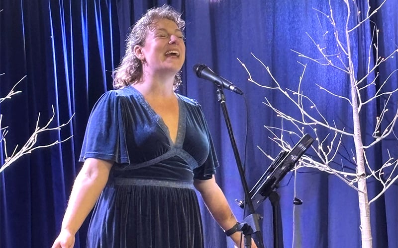 A woman in a blue dress sings passionately on stage, standing near a microphone and music stand, with white decorative tree branches and blue curtains in the background.